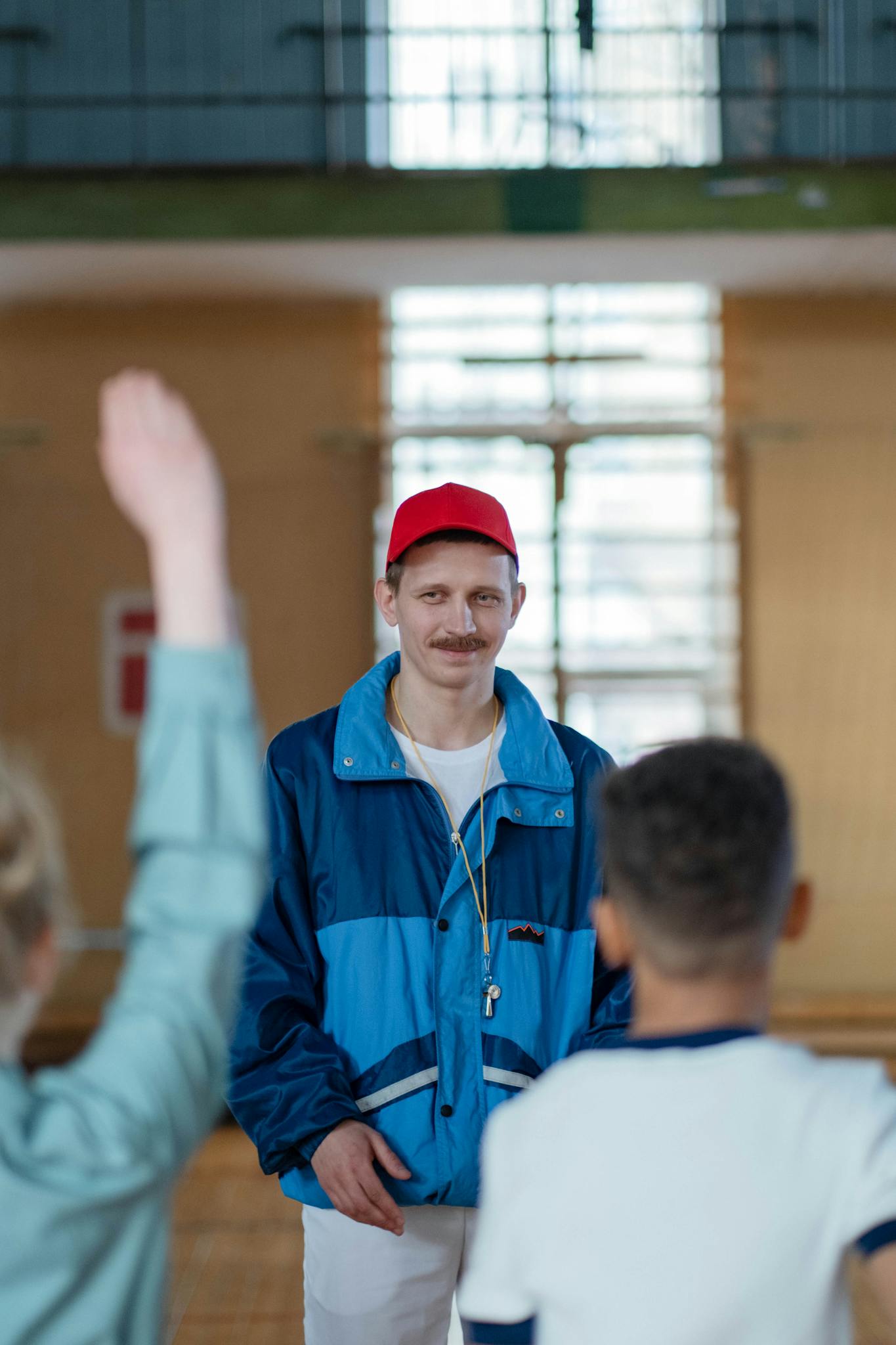 A sports coach in a red cap and blue jacket engages with students in a gym setting.